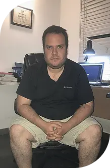 Hombre sentado en una oficina, con una expresión seria, frente a un escritorio y una computadora encendida.