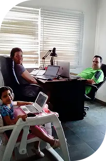 Tres personas trabajando en un espacio doméstico, dos adultos en escritorios y una niña usando una laptop en una silla.